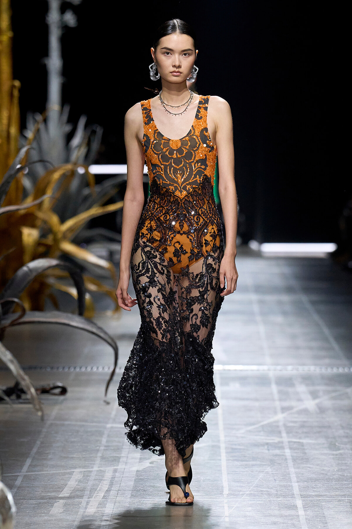 Model walks runway in an orange and black lace gown with intricate floral patterns and sheer black skirt, paired with bold earrings and sandals.