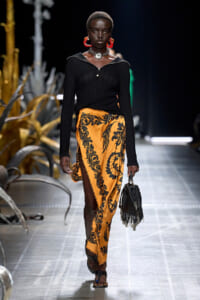 Model on a runway wearing a black knit top and a yellow-orange floral wrap skirt with a slit, paired with a black fringed handbag and bold red earrings.
