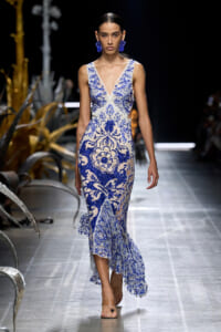 Model walking a runway in a blue-and-white lace gown with a plunging V-neck and scalloped hem with floral embroidery, worn with blue earrings.