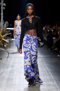 Model walks the runway in a black cropped hooded sweater with a lace-up front paired with blue-and-white floral wide-leg pants, carrying a black handbag on a fashion show runway.