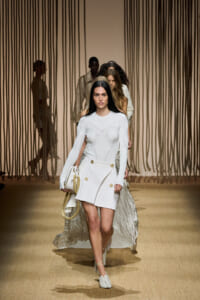 Model walks the runway in a white knit long-sleeve top and wrap mini skirt with gold buttons, holding a white bag with a rope handle.