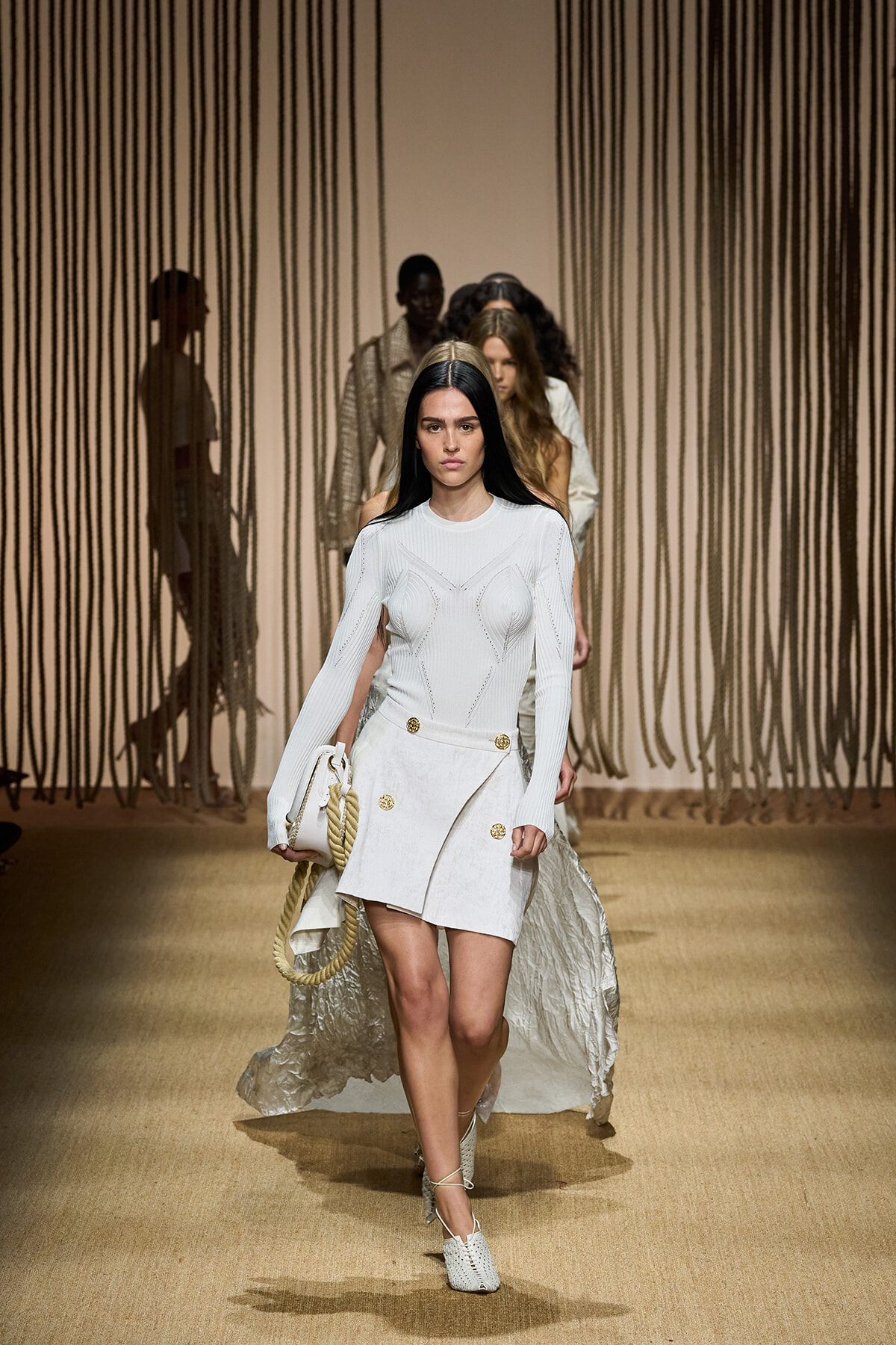 Model walks the runway in a white knit long-sleeve top and wrap mini skirt with gold buttons, holding a white bag with a rope handle.