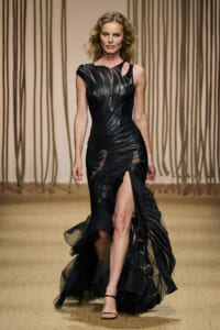 Model on a fashion runway wearing a black sculpted gown with sheer panels and a high slit, strappy sandals visible.