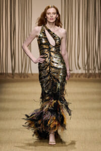 Model on a runway wearing a gold and black sequin one-shoulder gown with a feathered multicolor hem.