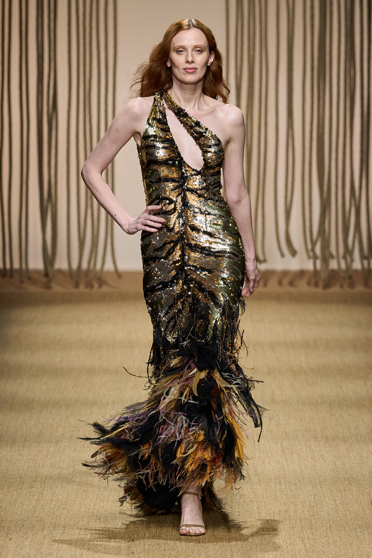 Model on a runway wearing a gold and black sequin one-shoulder gown with a feathered multicolor hem.