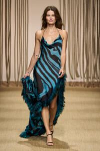 Model walking the runway in a teal and black zebra-print slip dress with teal feather trim along the hem; black strappy sandals.