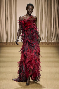 Model walking the runway in a burgundy feathered gown with bare shoulders and dark skin.
