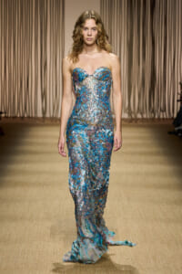 Model walking a runway in a strapless, turquoise-silver sequin gown with a flowing train against a beige backdrop.