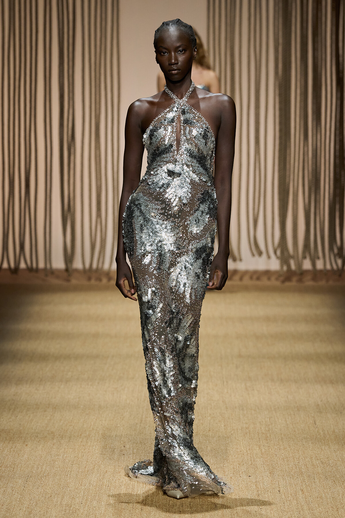 Runway model in a floor-length silver sequined halter gown with a crisscross neckline and subtle train on a fashion catwalk.