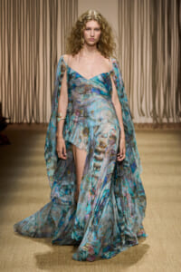 Model walks the runway wearing a teal-blue abstract-pattern gown with layered chiffon cape sleeves on a fashion show runway.