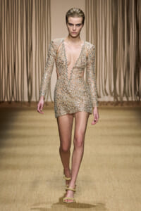 Model walking a runway in a nude-toned mini dress covered in silver beaded lattice, deep V neckline, long sheer sleeves, neutral backdrop.