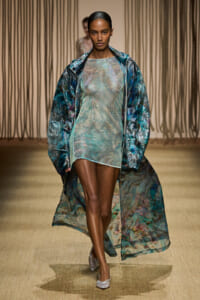 Model walks a runway in a shimmery, pastel-green mini dress paired with a long, translucent blue-green marbled coat.