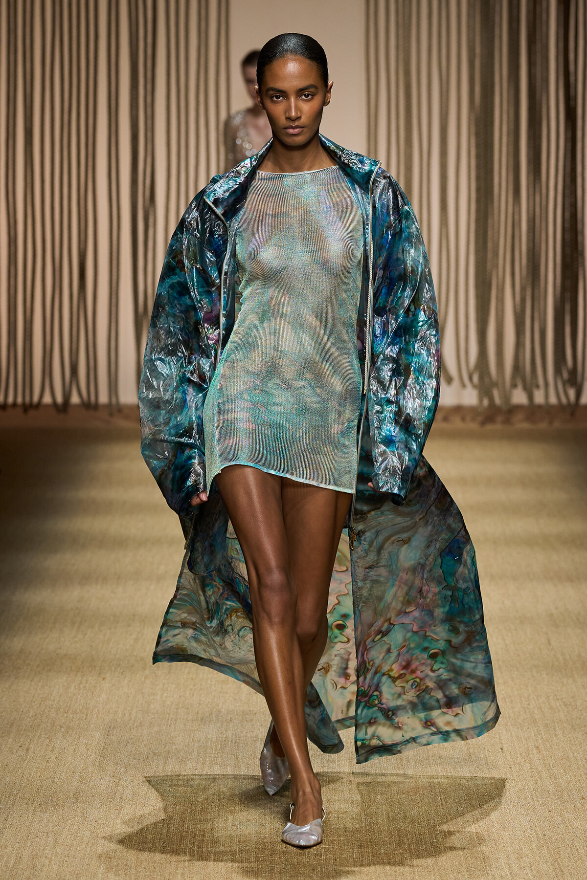 Model walks a runway in a shimmery, pastel-green mini dress paired with a long, translucent blue-green marbled coat.
