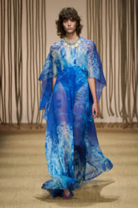 Model walks the runway in a flowing blue watercolor gown with a layered cape and statement necklace.
