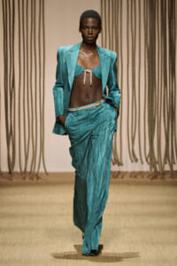 Model on a runway wearing a teal satin suit with an open blazer, a braided rope-tied bralette top, and high-waisted wide-leg pants; hands in pockets.