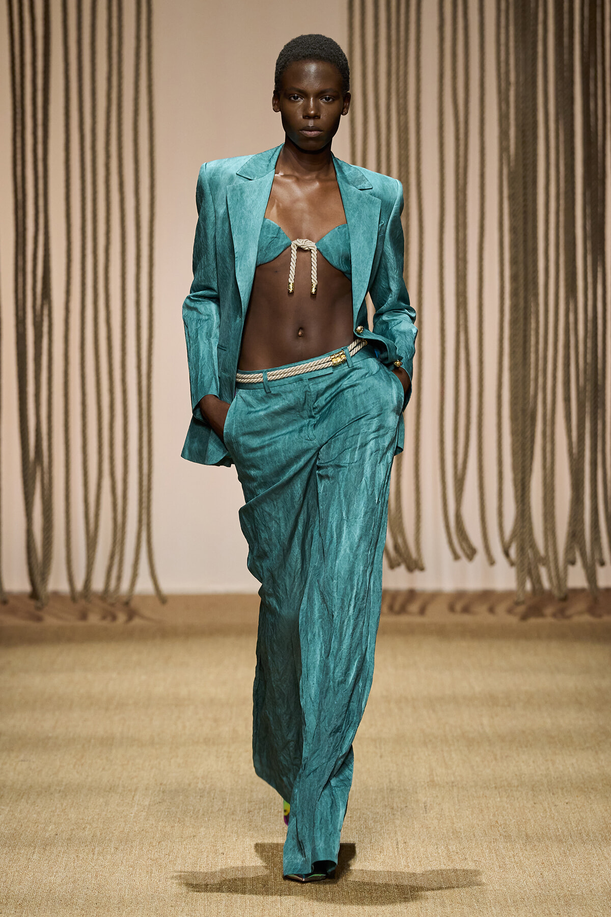 Model on a runway wearing a teal satin suit with an open blazer, a braided rope-tied bralette top, and high-waisted wide-leg pants; hands in pockets.