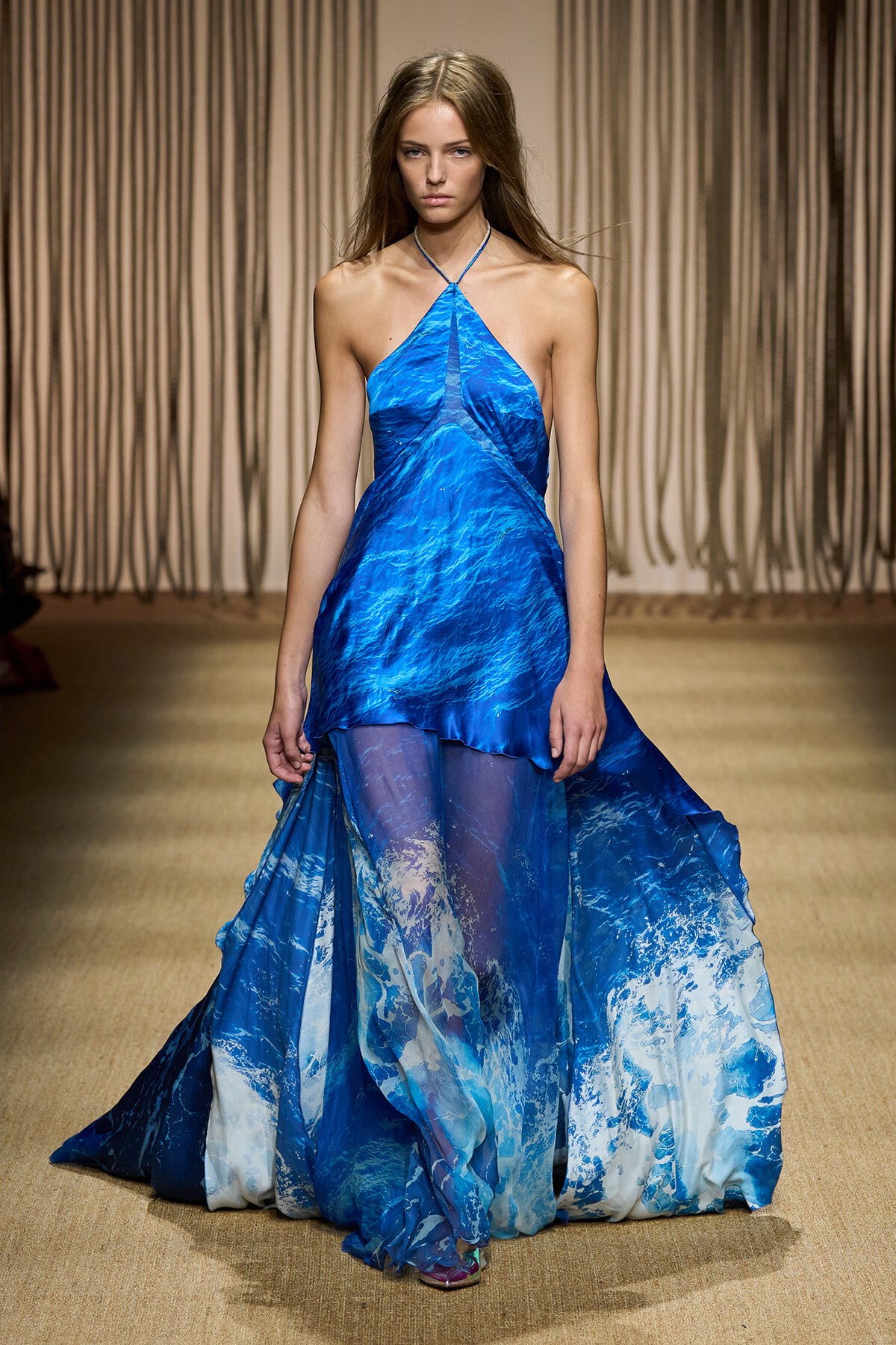 Model walks the runway in a vivid blue halter-neck gown with an ocean-wave print and a flowing high-low skirt.