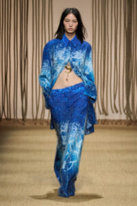Model walking runway in blue textured oversize shirt and matching pants, midriff exposed, gold pendant visible