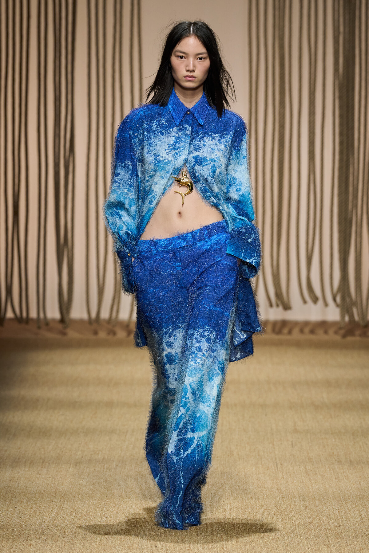 Model walking runway in blue textured oversize shirt and matching pants, midriff exposed, gold pendant visible