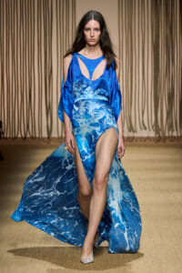 Model walks runway in a blue ocean-wave print gown with a high slit, sheer cape sleeves, and silver heels.