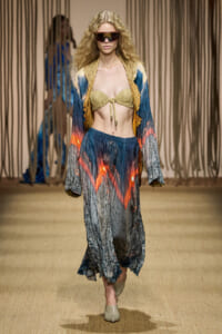 Model walking the runway in a colorful boho set: beige bikini top, blue-orange skirt, matching jacket, oversized visor sunglasses, and beige shoes.