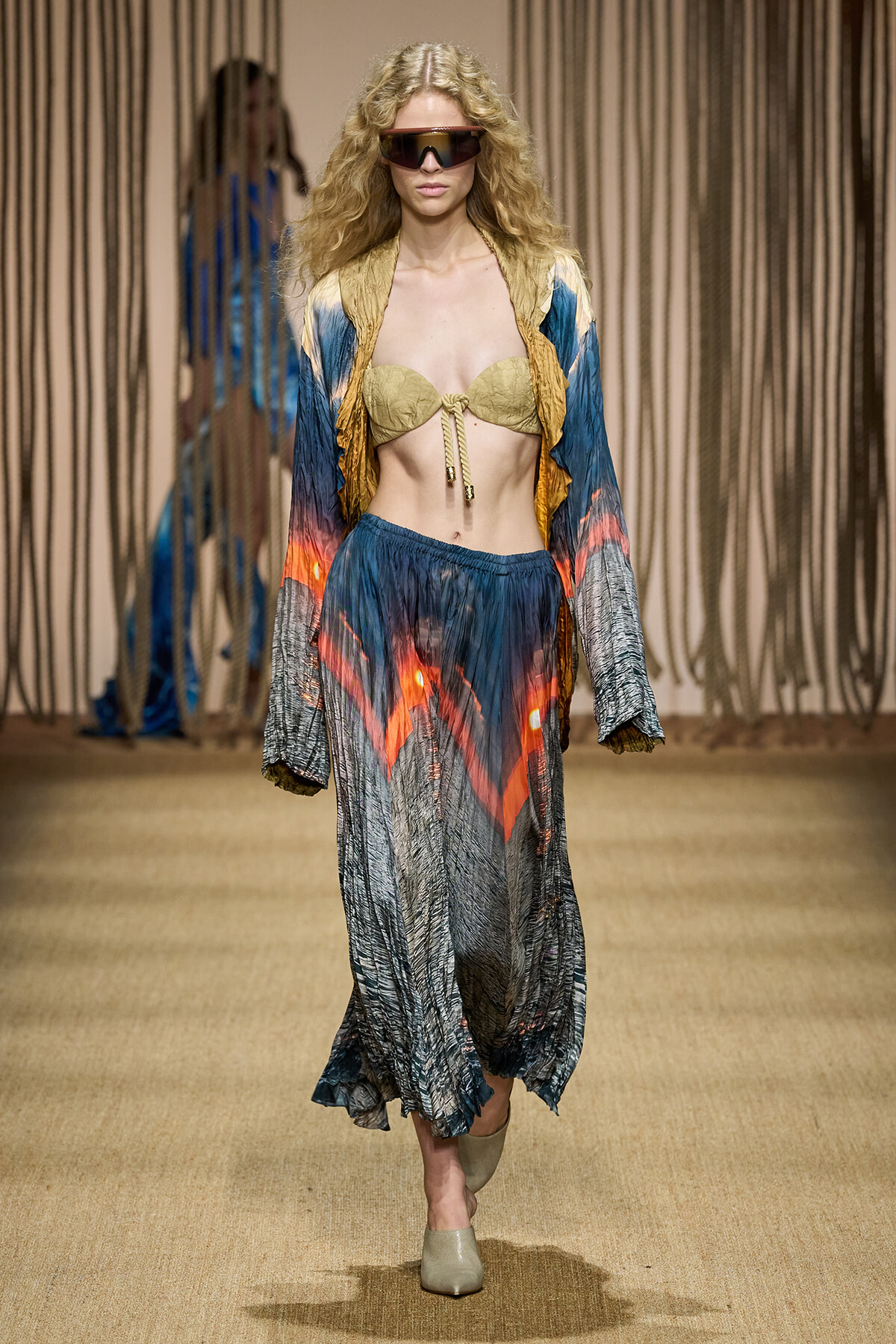 Model walking the runway in a colorful boho set: beige bikini top, blue-orange skirt, matching jacket, oversized visor sunglasses, and beige shoes.