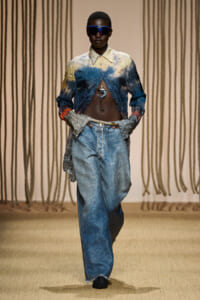 Model on a runway wearing a blue-yellow gradient cropped jacket with exposed midriff and baggy jeans with futuristic sunglasses.