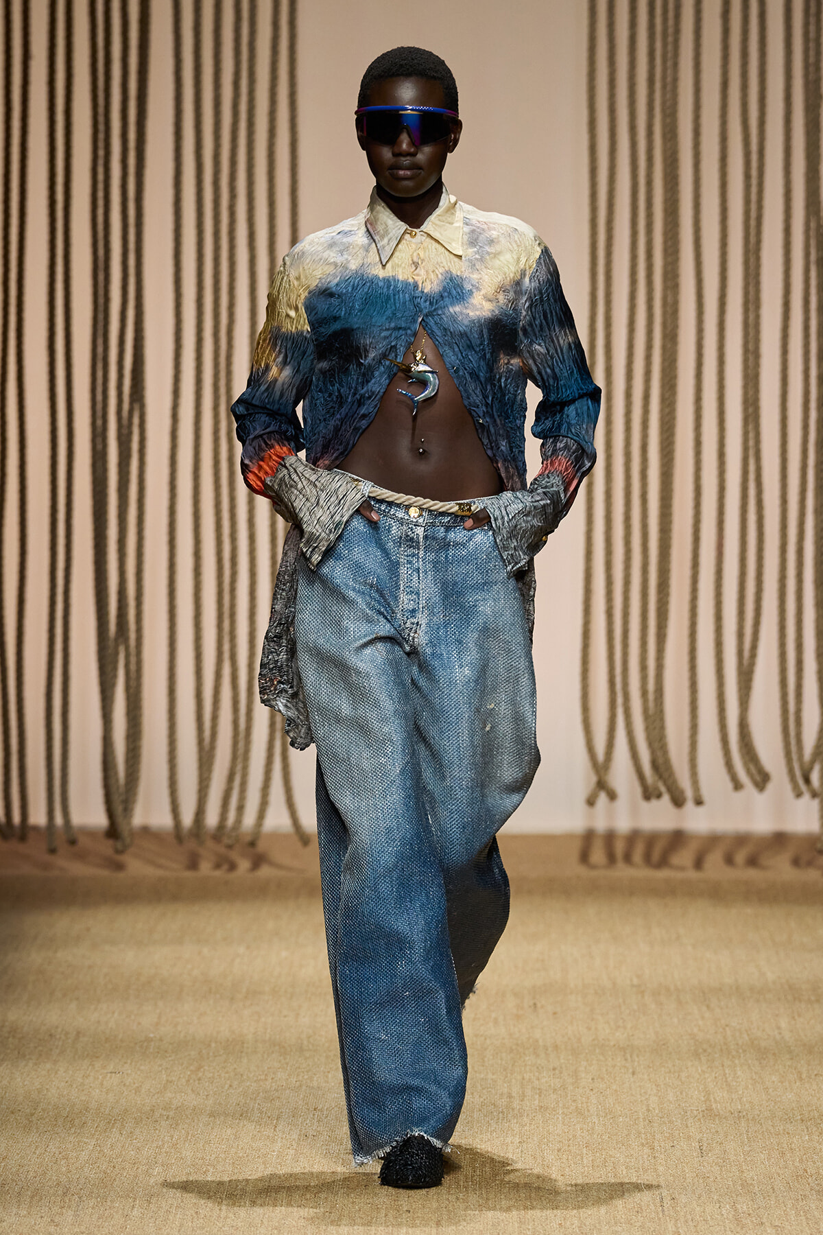 Model on a runway wearing a blue-yellow gradient cropped jacket with exposed midriff and baggy jeans with futuristic sunglasses.