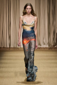 Model walking a runway in a long, gradient slip dress with sunset hues and wave-like patterns.