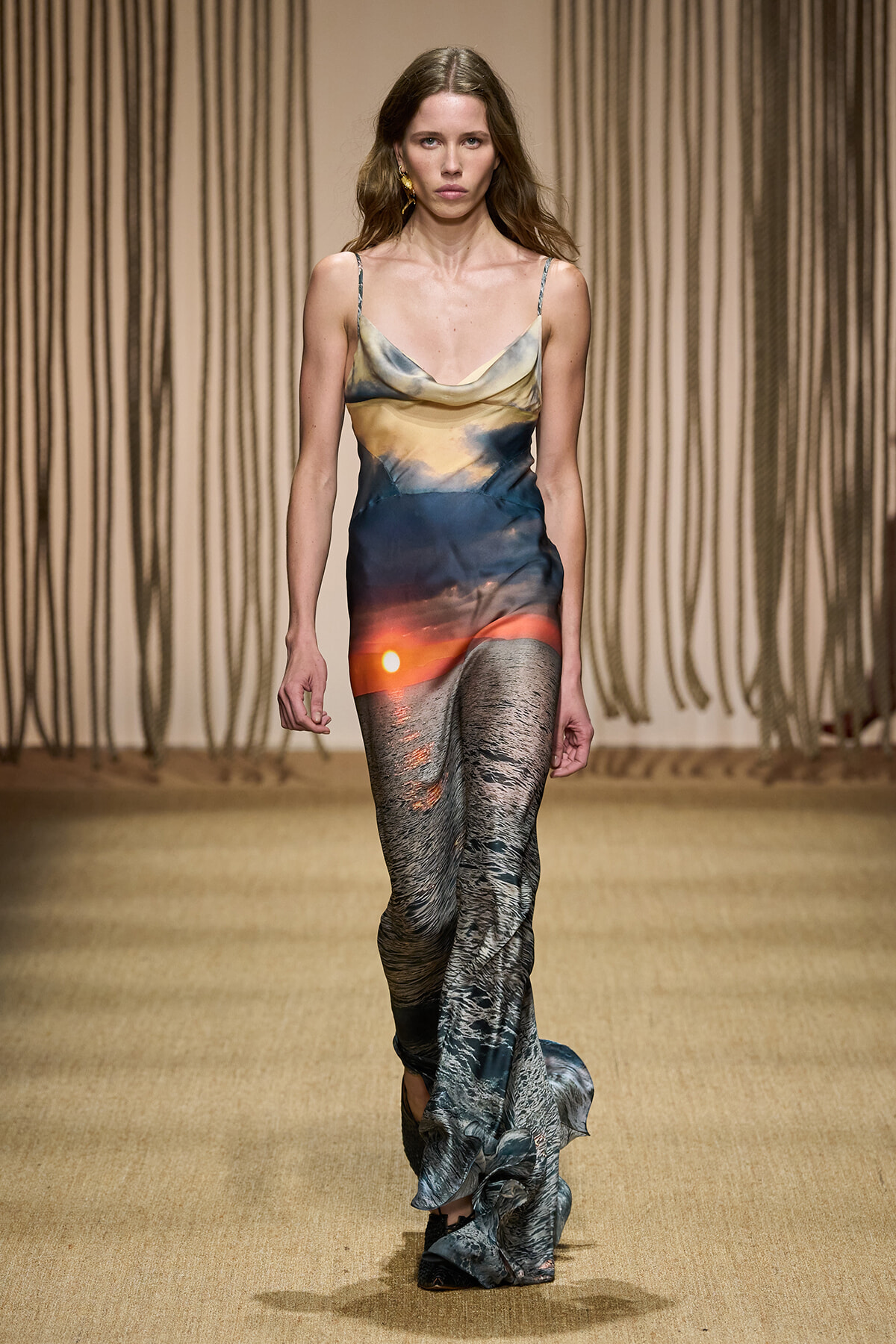 Model walking a runway in a long, gradient slip dress with sunset hues and wave-like patterns.
