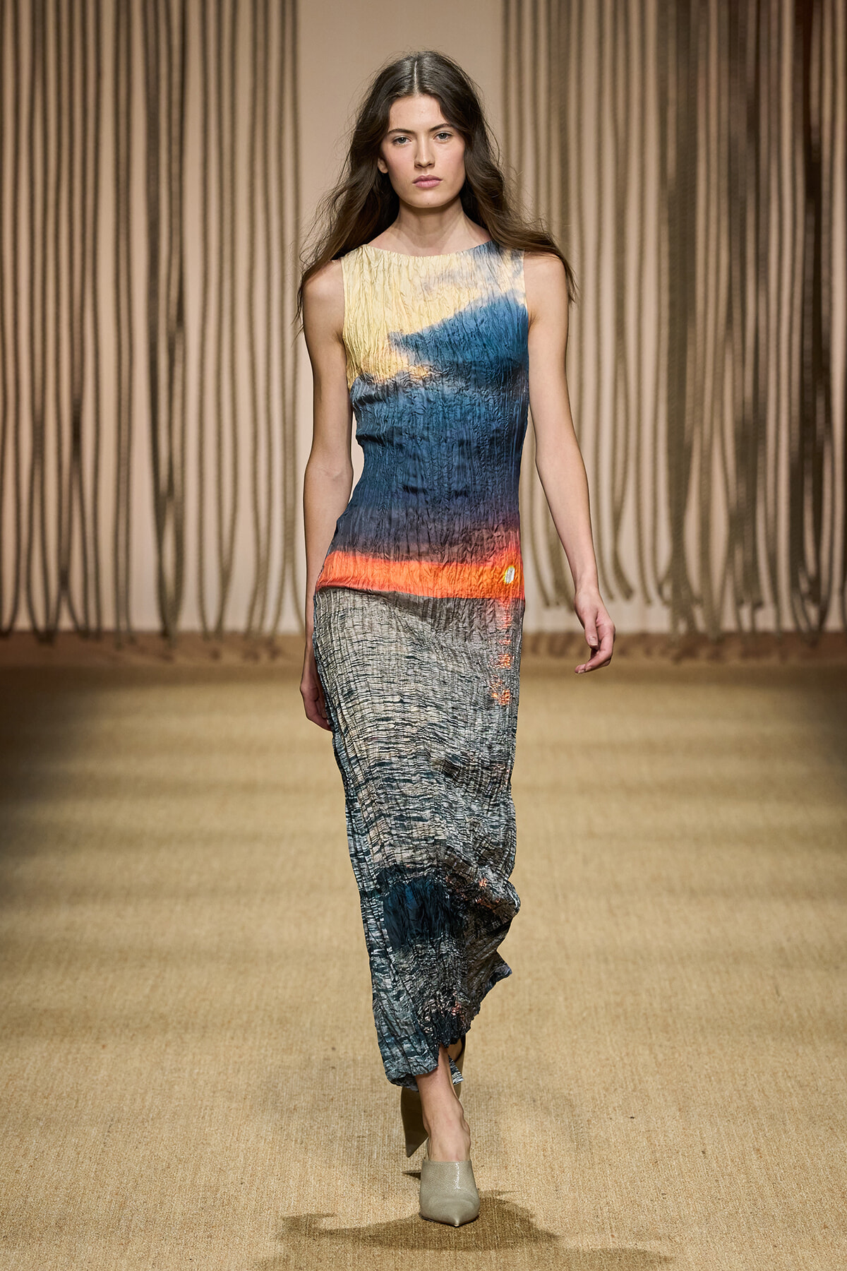 Model walks the runway in a sleeveless, crinkled maxi dress with gradient yellow, blue, orange, and gray tones, paired with beige heels.