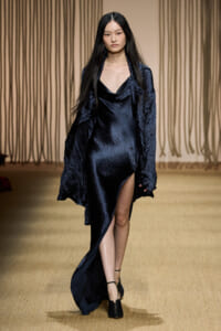 Female model on a fashion runway wearing a navy velvet draped dress with a high slit and black ankle-strap heels, long dark hair flowing.
