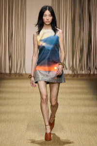 Model walking a runway in a sleeveless gradient mini dress that shifts from pale yellow to blue to orange, with brown heeled sandals and gold bracelets.
