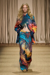Runway model walks center stage in a vibrant, abstract-print matching shirt and wide-leg pants set with a midriff showing knot detail.