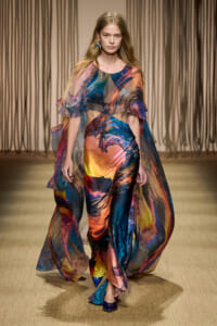 Model walks runway in a vibrant, multicolored abstract-print dress with a sheer cape-like overlay and matching blue shoes, on a beige runway backdrop.