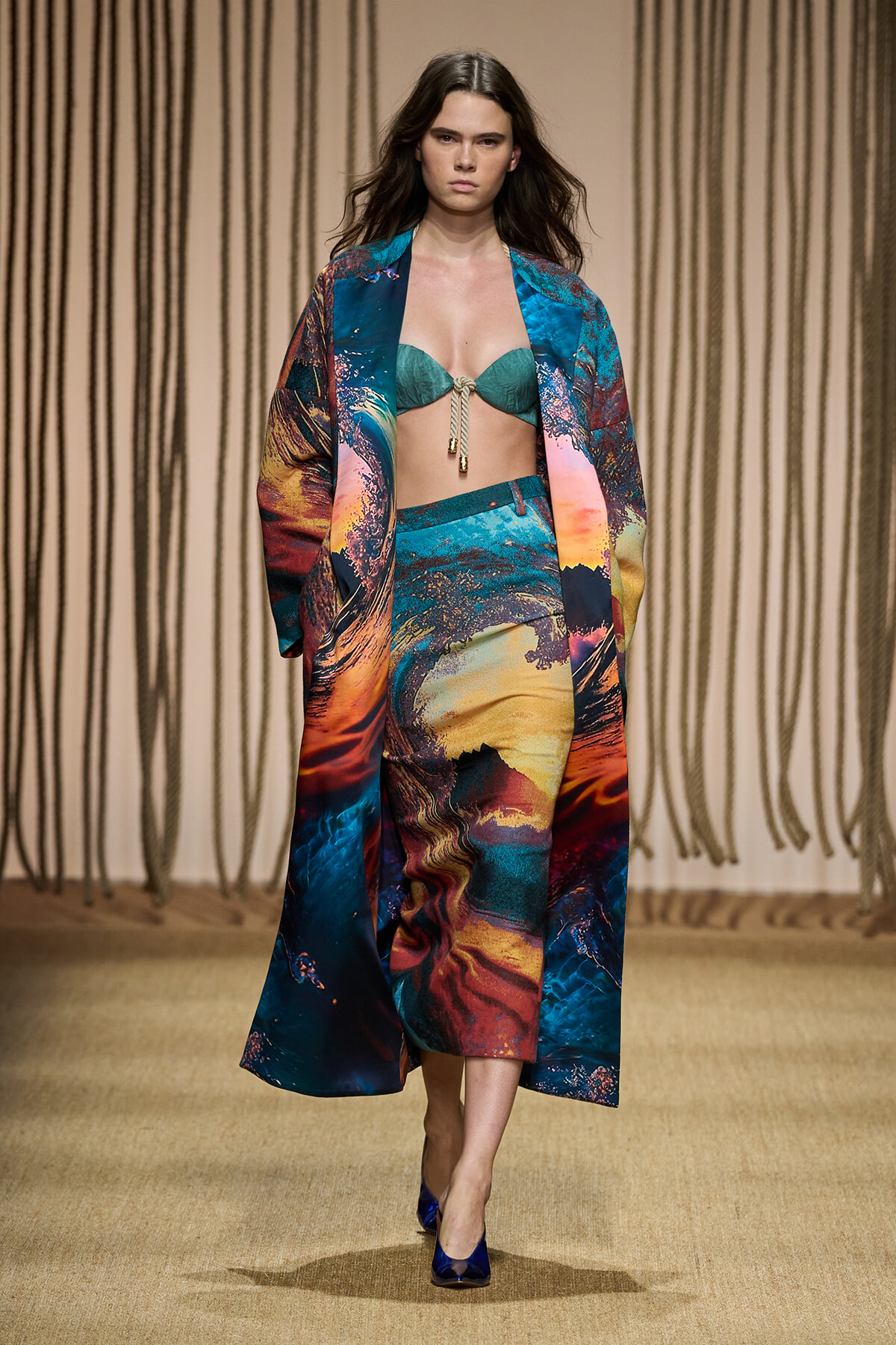 Runway model in a bold turquoise, orange, and blue abstract-print coat and matching skirt, with a teal bikini top and blue heels.