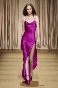 Model on a runway wearing a magenta satin draped halter gown with a thigh‑high slit and matching heels on a fashion show catwalk.