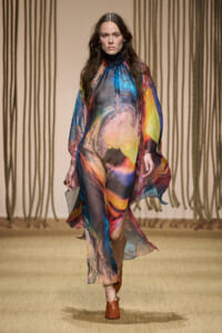 Model walks the runway in a loose, rainbow-hued abstract-print chiffon dress with long sleeves and a flowing silhouette on a beige catwalk.