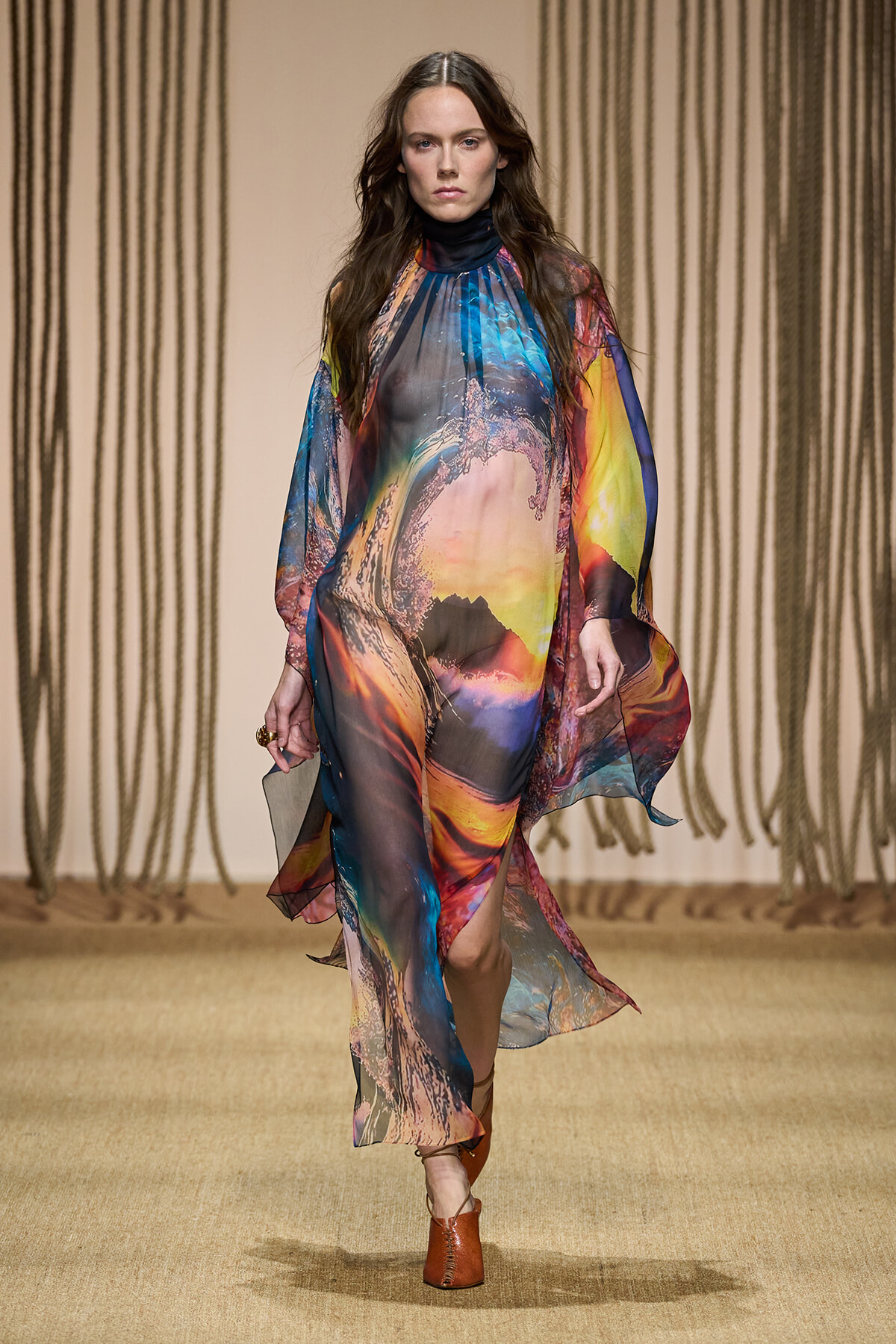 Model walks the runway in a loose, rainbow-hued abstract-print chiffon dress with long sleeves and a flowing silhouette on a beige catwalk.