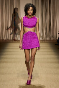 Model walking a runway in a bright pink textured two-piece dress with a midriff cutout tied by a rope, matching pink heels, and bold jewelry.