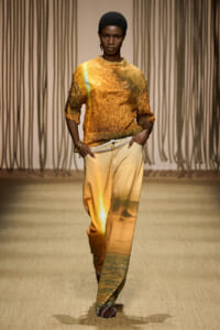 Model walking a runway in an orange-gold crinkled top and tan-to-gold gradient wide-leg pants with a rope belt.