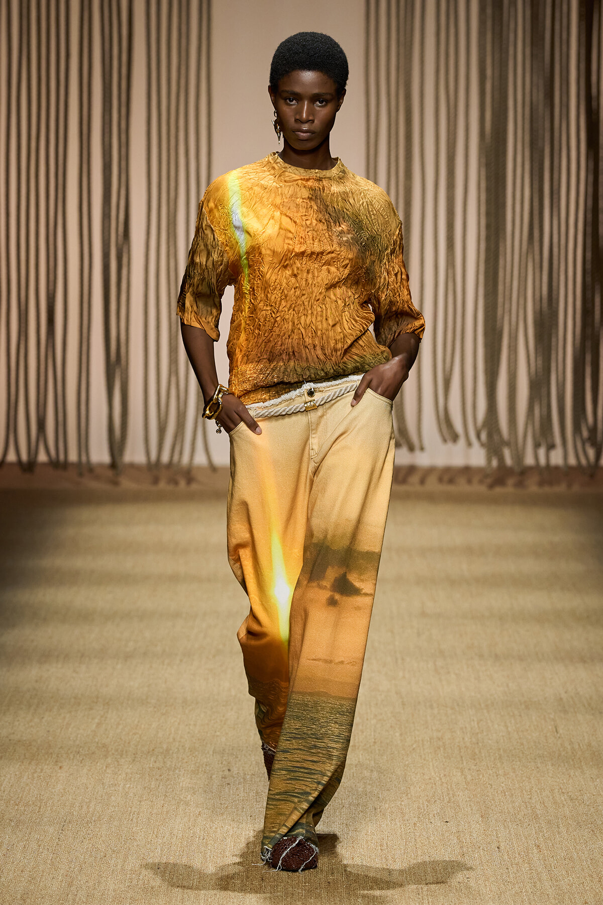 Model walking a runway in an orange-gold crinkled top and tan-to-gold gradient wide-leg pants with a rope belt.