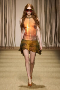 Model walking a runway in a metallic orange sleeveless top, layered bronze-gold skirt, and futuristic green-t rim sunglasses; long red hair down.