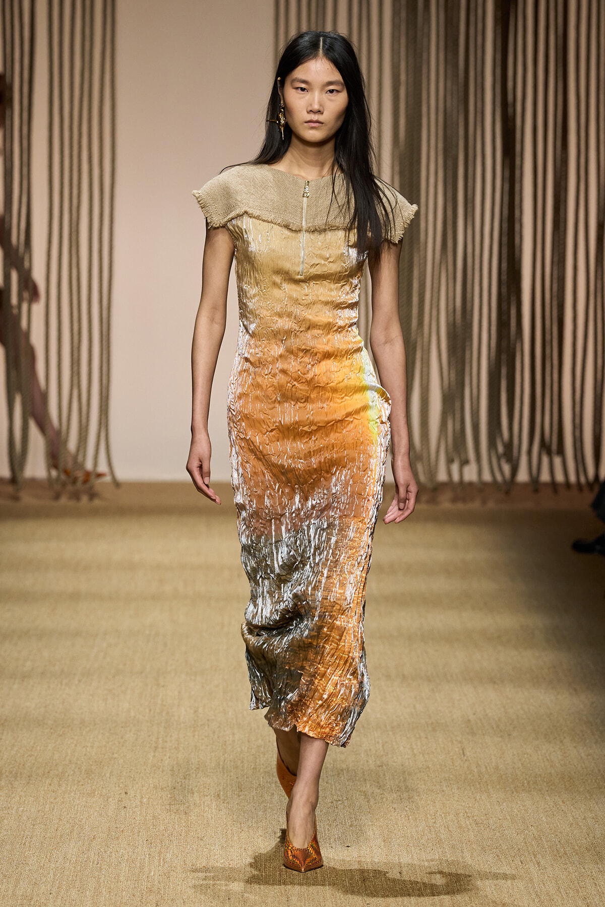 Model walks the runway wearing a crinkled metallic gradient dress from beige to orange with a fringed collar and orange heels.
