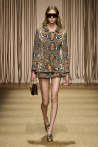 Model walking a runway in a brown snakeskin-print tailored blazer and matching layered shorts, with oversized sunglasses and snakeskin heels, carrying a small crocodile-pattern handbag.