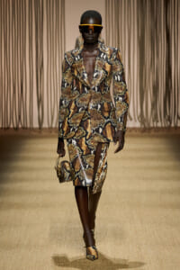 Runway model in a brown snakeskin-print suit with a bold blazer, matching skirt, orange visor sunglasses, and a small snakeskin handbag.