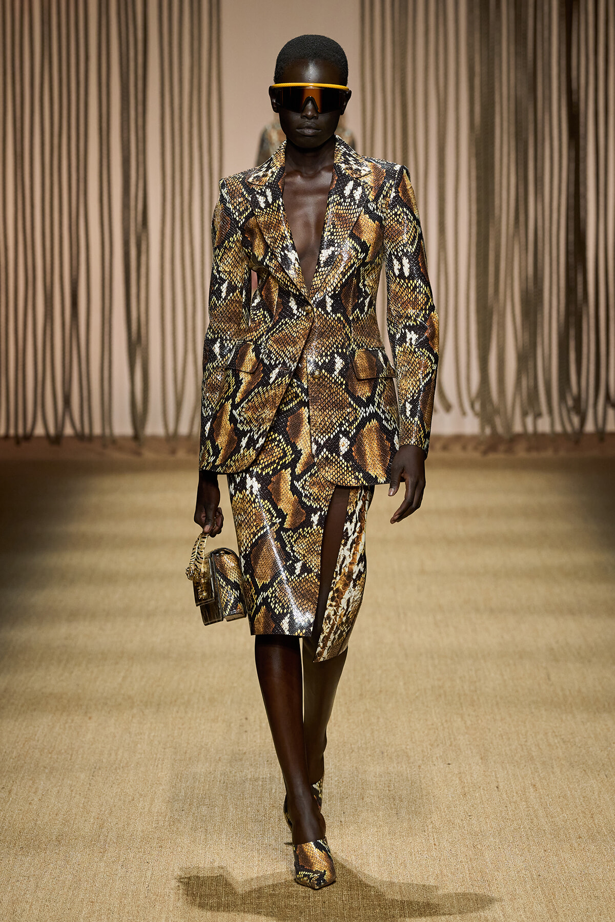 Runway model in a brown snakeskin-print suit with a bold blazer, matching skirt, orange visor sunglasses, and a small snakeskin handbag.