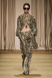 Runway model in a snakeskin-print blazer and wide-leg pants, midriff exposed, wearing futuristic sunglasses.