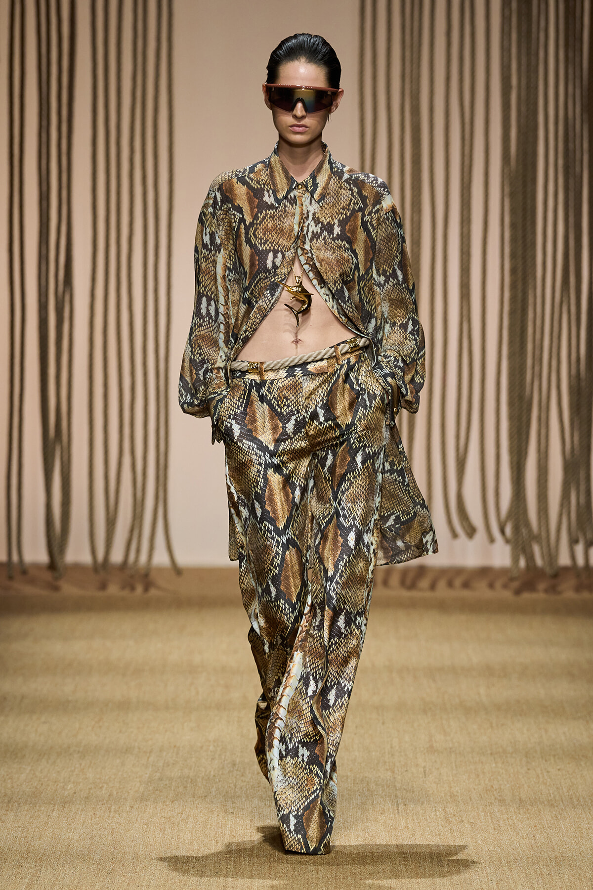 Runway model in a snakeskin-print blazer and wide-leg pants, midriff exposed, wearing futuristic sunglasses.