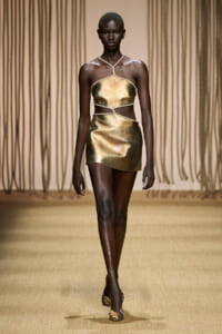 Model walks the runway in a gold metallic two-piece outfit with rope halter and wrap details on a fashion show runway.